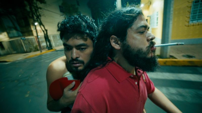 Dos hombres en una motocicleta en movimiento de noche por la calle; el conductor tiene barba y un cigarrillo encendido en la boca mientras el pasajero lo abraza tenso por la espalda.