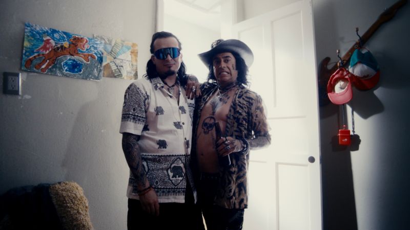 Dos hombres con vestimenta estrafalaria, tatuajes visibles, sombreros y lentes oscuros posando juntos en el marco de la puerta de una habitación.