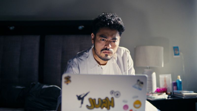 Un hombre sentado en la cama frente a una computadora portátil con calcomanías, mirando fijamente la pantalla con expresión de preocupación y concentración en una habitación penumbrosa.