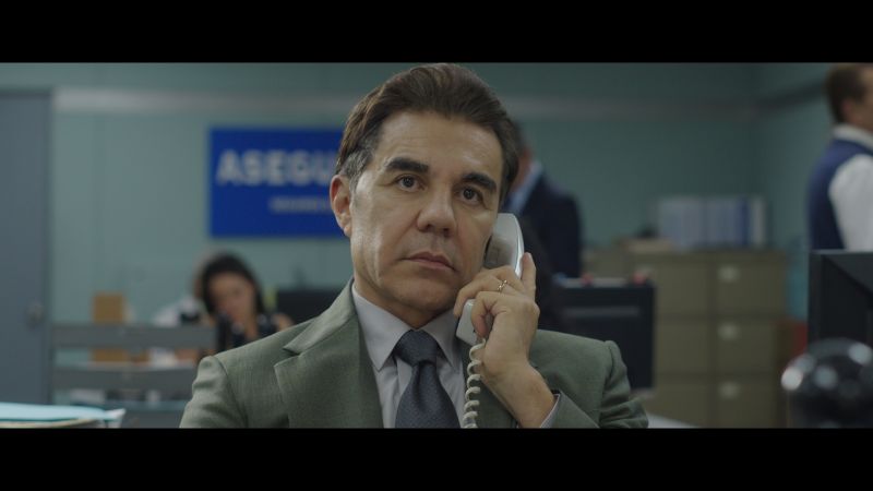 Adrián Uribe como Juan Pablo en su oficina de seguros Primer plano de Juan Pablo González (Adrián Uribe) hablando por teléfono de oficina con expresión seria y pensativa en su escritorio de trabajo.