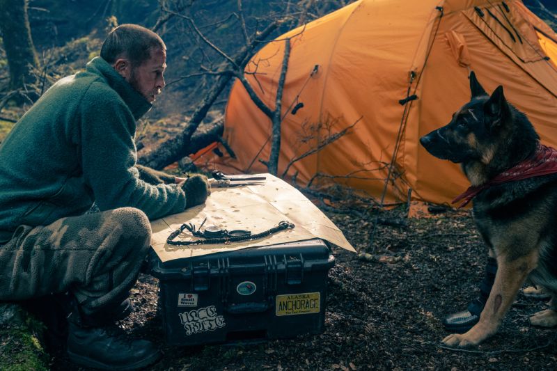 Brad Pitt agachado junto a un estuche táctico revisando un mapa frente a su perro pastor alemán cerca de una tienda de campaña naranja.