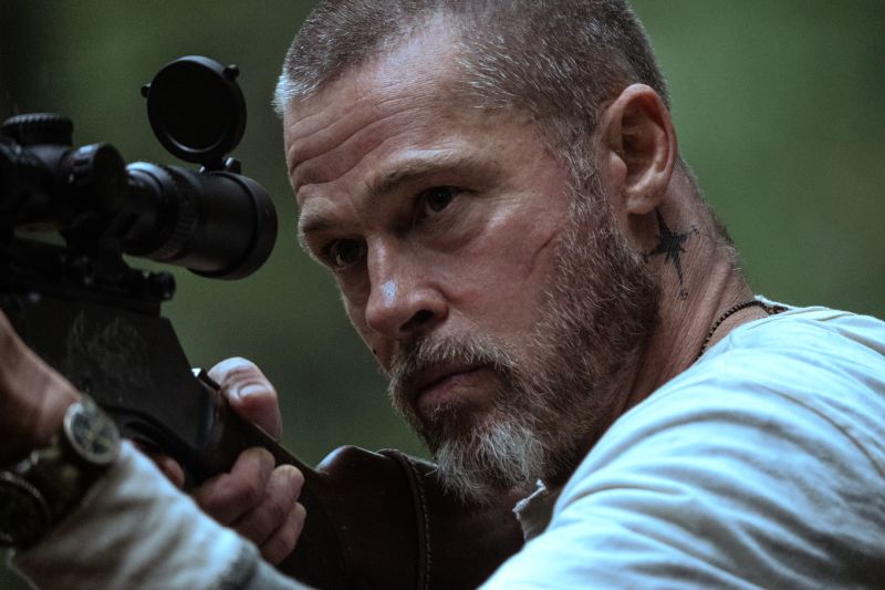 Primer plano del rostro de Brad Pitt apuntando con un rifle con mira telescópica entre la vegetación.