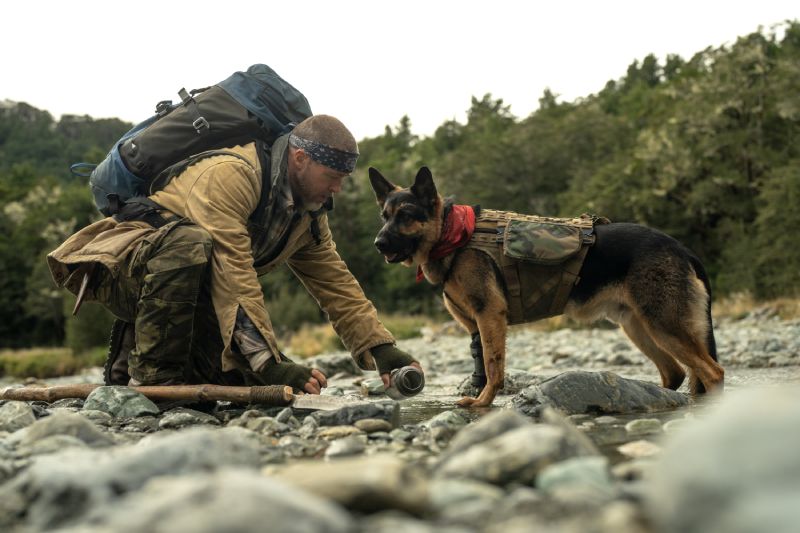 Brad Pitt agachado en la orilla de un río llenando una cantimplora junto a su perro Odín que bebe agua a su lado.