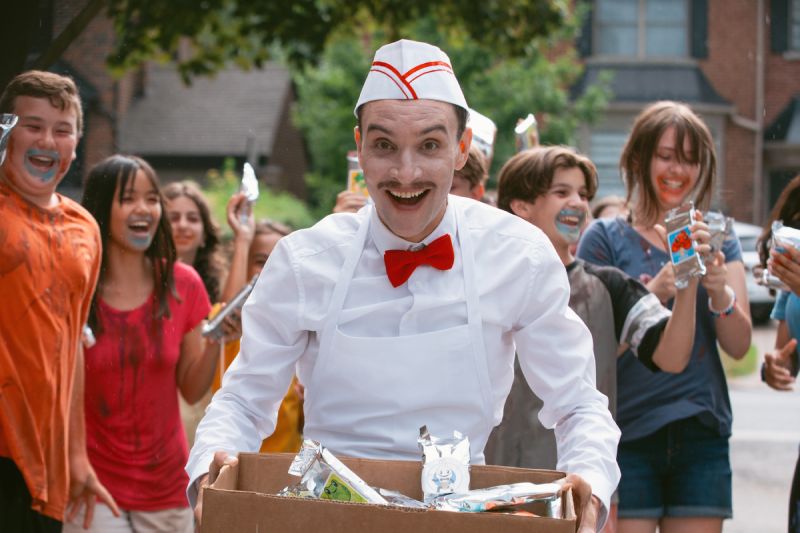 El actor caracterizado como el alegre y siniestro heladero sonríe eufóricamente mientras viste un uniforme blanco con moño rojo, cargando una caja de golosinas rodeado de niños entusiasmados.