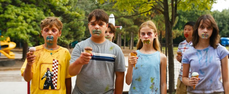 Cuatro niños parados en línea en un parque sosteniendo conos de helado, con miradas perdidas y expresiones catatónicas, con manchones coloridos y oscuros alrededor de sus bocas.
