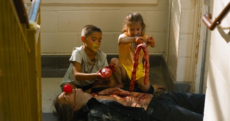 Dos niños pequeños sentados en el suelo jugando violentamente con las vísceras e intestinos de un hombre sin vida tirado a su lado con una nariz de payaso.