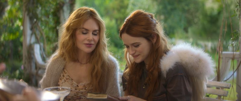 Gillian Owens y la nueva generación de la familia Owens Nicole Kidman en suéter beige junto a Joey King leyendo sonrientes un libro antiguo sobre una mesa en el jardín rodeadas de vegetación.