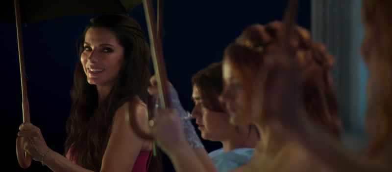 Sally Owens en una escena festiva nocturna Sandra Bullock sonriendo bajo un paraguas abierto durante una celebración nocturna rodeada por miembros de su familia vestidas de gala.
