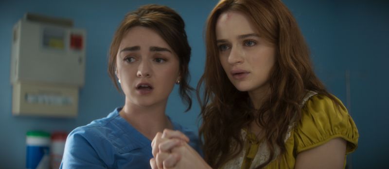 Maisie Williams y Joey King en un momento de tensión dramática Maisie Williams vestida con uniforme médico azul sosteniendo con preocupación la mano de Joey King en una habitación de hospital.