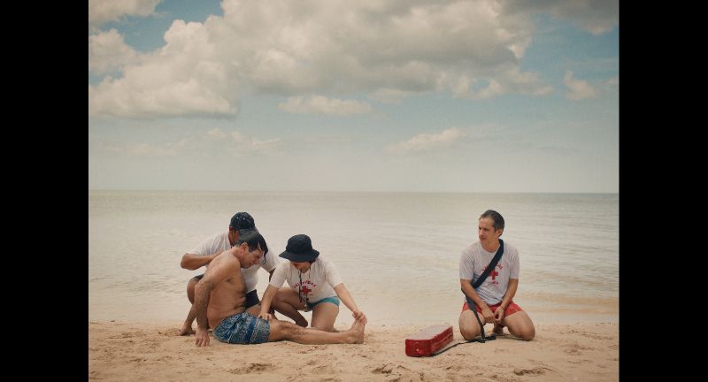 Fotograma de la película mostrando a un hombre rescatado en la playa siendo atendido por paramédicos y un salvavidas sentado al lado sobre la arena.