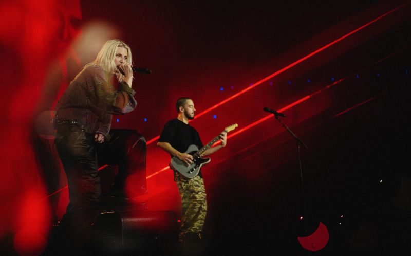 Emily Armstrong en vivo en Linkin Park: Unshatter Emily Armstrong cantando en vivo en un escenario iluminado con luces rojas intensas durante una presentación de Linkin Park.
