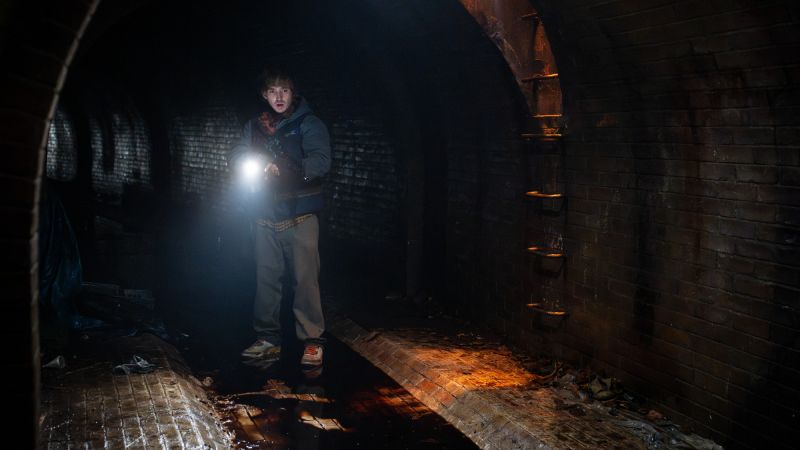 Bryan explora los túneles subterráneos en Resident Evil: Noche Cero Bryan (Austin Abrams) parado en medio de un túnel o drenaje subterráneo oscuro de ladrillo, sosteniendo una linterna encendida que ilumina el ambiente húmedo y sombrío.