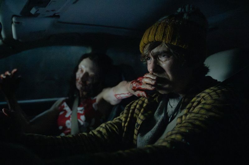 Enfrentamiento en el interior de un automóvil en Resident Evil: Noche Cero Bryan aterrorizado dentro de un vehículo de noche con el cinturón puesto, cubriéndose la boca ensangrentada mientras una figura femenina infectada se abalanza desde el asiento trasero.