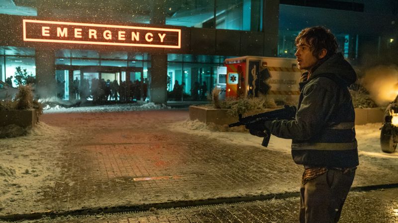 Escena en el exterior del hospital de emergencias en Resident Evil: Noche Cero Bryan parado afuera de la entrada iluminada de emergencias de un hospital en medio de la nieve, sosteniendo un arma subametralladora mientras mira hacia los lados alerta.