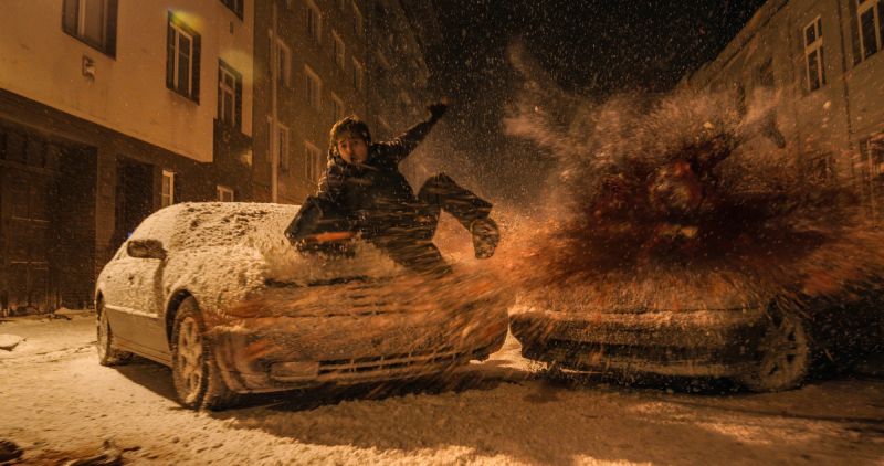 Impacto y acción en la calle nevada en Resident Evil: Noche Cero Escena de acción nocturna en la calle cubierta de nieve donde Bryan sale disparado e impacta sobre el cofre de un automóvil durante una violenta explosión o colisión.