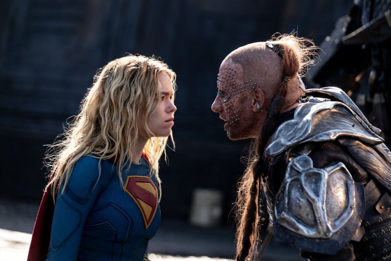Confrontación directa en Supergirl Plano medio de un tenso enfrentamiento cara a cara entre Supergirl (Milly Alcock) y un temible cazador de recompensas alienígena con implantes metálicos en el rostro.