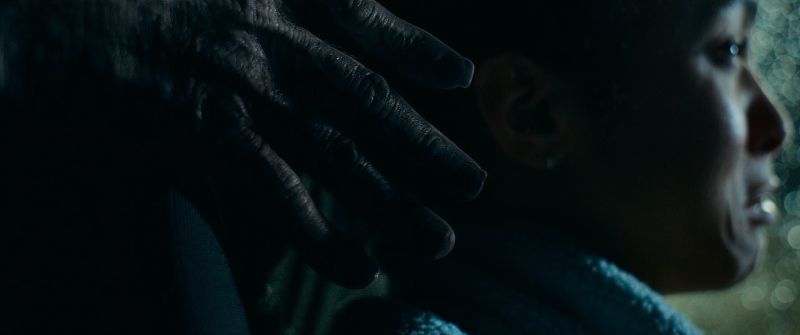 Mano oscura acercándose al hombro de una persona en una escena de tensión con iluminación tenue.