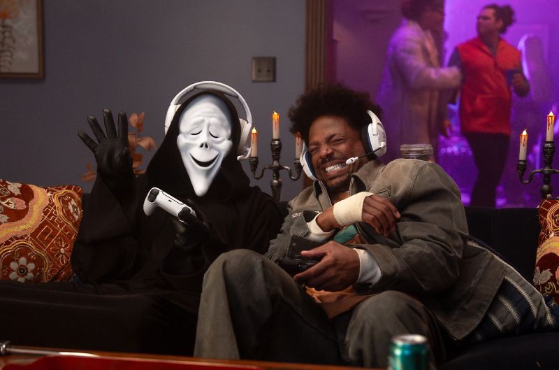 Scary Movie Terrorificamente Incorrecta escena de comedia en formato digital Un hombre sentado en un sillón junto a un personaje con la túnica negra y máscara sonriente de Ghostface. Ambos sostienen mandos de consola de videojuegos y llevan auriculares blancos con micrófono integrados, riendo en una habitación decorada con velas sobre candelabros.