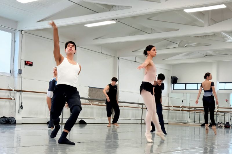 Ensayos de Grafías por la Compañía Nacional de Danza Fotografía de ensayo en salón de danza iluminado donde varios bailarines y bailarinas de la Compañía Nacional de Danza realizan movimientos contemporáneos y clásicos; en primer plano un bailarín con camiseta blanca extiende el brazo hacia arriba mientras una bailarina en puntas realiza una postura grácil.