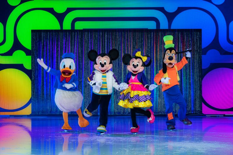 Disney On Ice — Anfitriones clásicos Mickey, Minnie, Donald y Goofy posan juntos en un escenario iluminado con patrones multicolores.