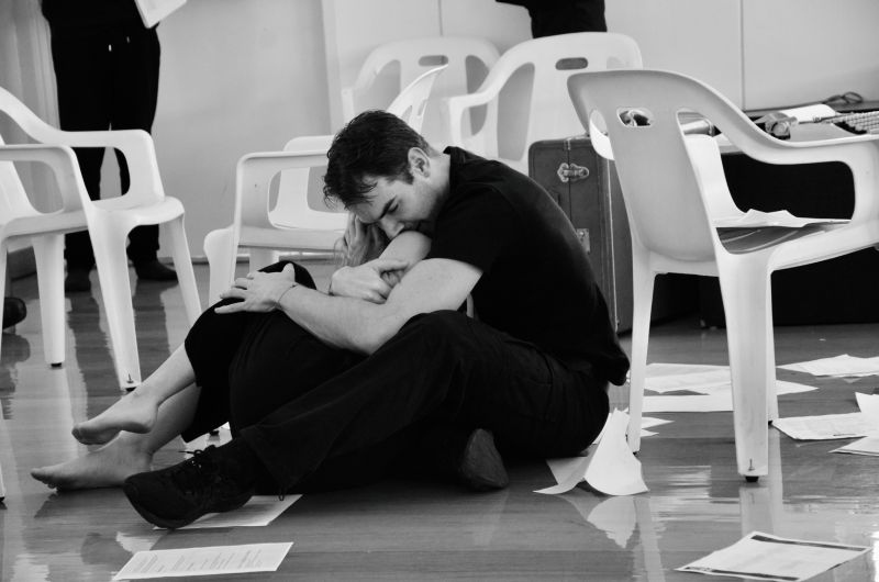Ojos de atardecer – Ensayo escénico Dos personas sentadas en el suelo se abrazan en un espacio interior rodeado de hojas sueltas y sillas.