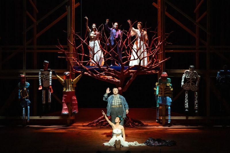 El último sueño de Frida y Diego – Producción del Met Escena teatral con un árbol escultórico rojo en el centro, rodeado de intérpretes y figuras humanas estilizadas en un escenario iluminado.