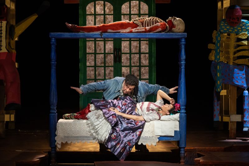Escena teatral con cama, figuras esqueléticas y estética inspirada en el Mictlán.