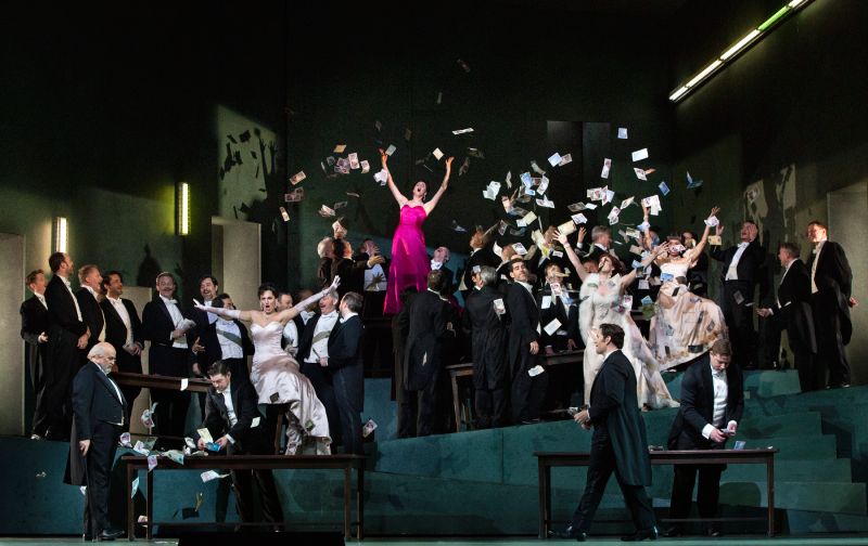 Montaje de la ópera Manon en el Met de Nueva York Fotografía de la puesta en escena de la ópera Manon sobre el escenario del Metropolitan Opera House. En el centro, una solista con vestido de gala rosa celebra mientras billetes de papel caen sobre el elenco distribuido en una escenografía inclinada de tonos verdes y elegantes trajes de etiqueta.