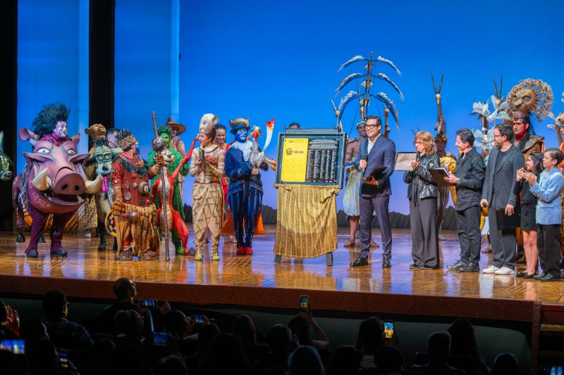 Develación de la placa por 500 representaciones de El Rey León Fotografía del elenco del musical El Rey León en el escenario junto a ejecutivos y creativos durante la develación de la placa conmemorativa. Al centro se observa la placa colocada sobre un atril dorado mientras los intérpretes caracterizados con sus vestuarios y máscaras aplauden.