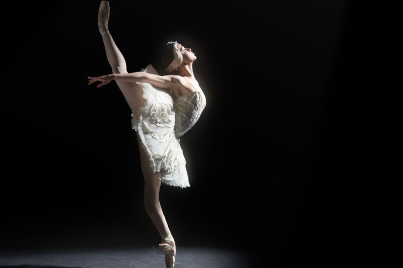Presentación de El lago de los cisnes por la Compañía Nacional de Danza Bailarina caracterizada como el personaje del cisne en una pose de ballet clásico, sobre un fondo oscuro iluminado por un haz de luz cenital. Viste tutú blanco detallado y mantiene una pierna elevada en extensión vertical sobre punta.