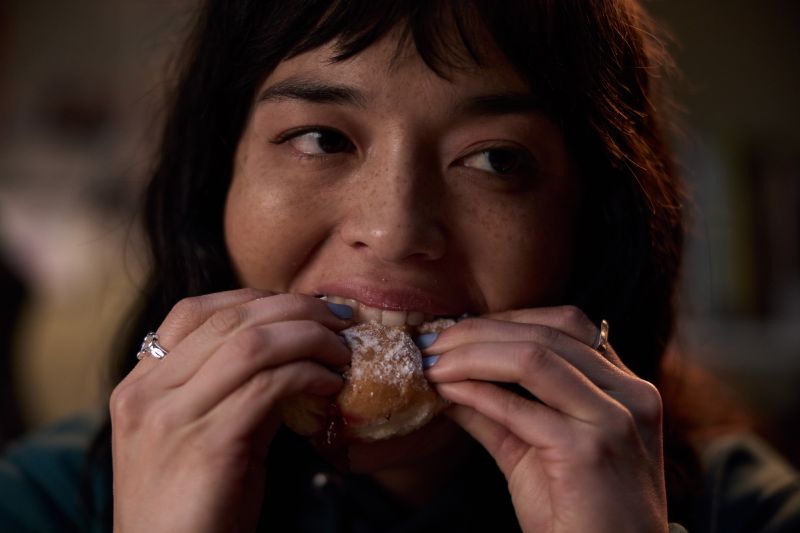 Hanna en una escena de Insaciable Plano detalle de Hanna mirando fijamente hacia el frente mientras muerde con ansiedad un pan dulce con mermelada y azúcar glas sosteniéndolo con ambas manos.