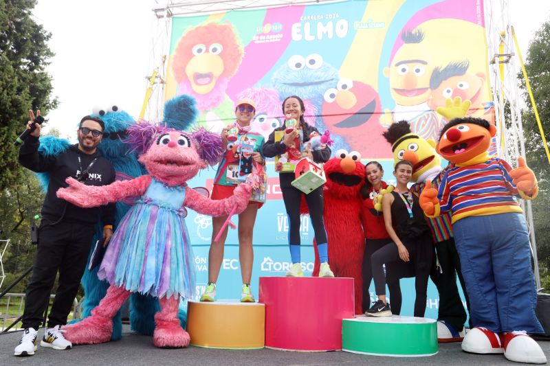 Ganadoras y personajes de Plaza Sésamo en el podio de la Carrera Elmo 2026 Fotografía en el podio de premiación donde dos corredoras posan con sus trofeos acompañadas por las botargas de Abby Cadabby, Elmo, Beto, Enrique y los presentadores del evento.