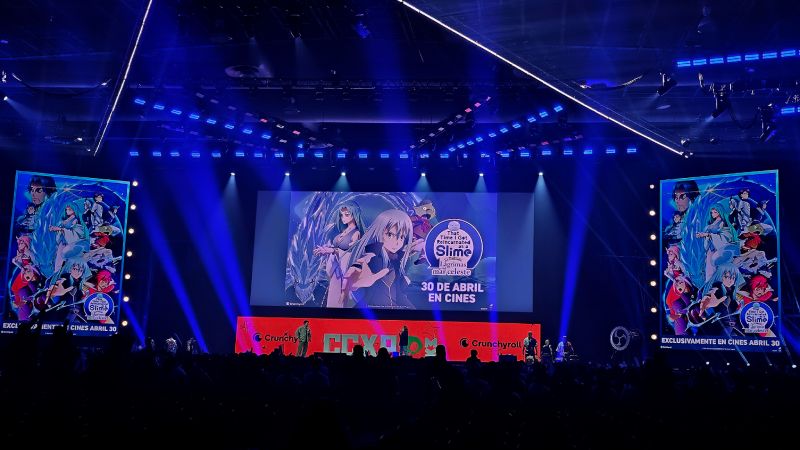 Escenario iluminado en tonos azules y morados con una pantalla gigante que muestra arte promocional de That Time I Got Reincarnated as a Slime: The Movie – Scarlet Bond, acompañado de banners laterales y público asistente.