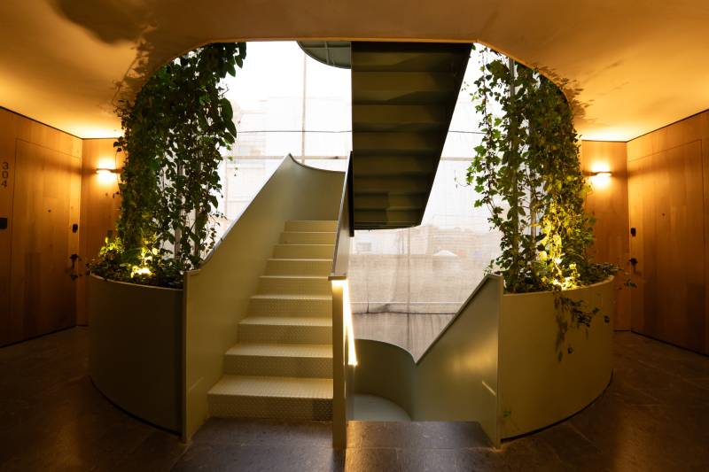 Escalera central de diseño moderno rodeada de jardineras curvas con vegetación alta, iluminación cálida y acabados en madera que enmarcan un ambiente interior armónico.
