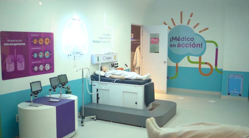 Sala educativa con una cama hospitalaria, un maniquí médico y proyecciones luminosas que explican funciones del cuerpo humano en un entorno interactivo.