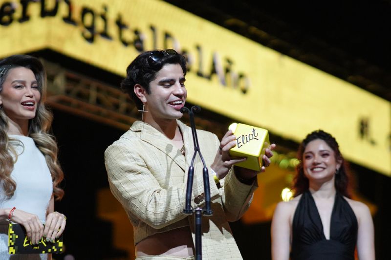 El creador de contenido Aaron Mercury en el escenario de los Eliot Awards 2026 sosteniendo con ambas manos la estatuilla dorada con forma de cubo que lleva la leyenda Eliot. Viste un saco corto en tono crema de cuello estilizado, mientras al fondo la conductora Galilea Montijo sonríe luciendo un vestido blanco de gala frente a las pantallas gigantes amarillas del evento.