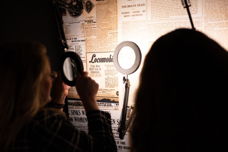 Consulta de Archivos Históricos en la Exposición Visitantes utilizando lupas de aumento para leer titulares y notas impresas sobre portadas de periódicos históricos dedicados al hundimiento del Titanic.