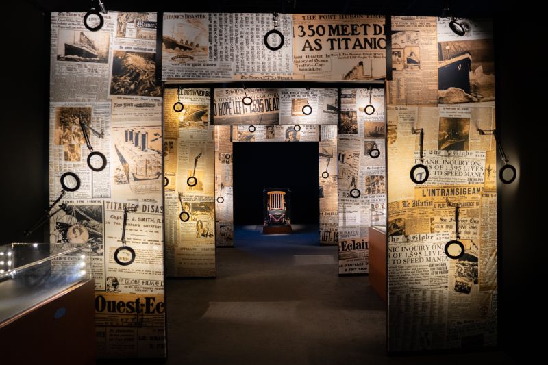 Recorrido por la Hemeroteca y Salas Temáticas Vista de un pasillo expositivo con paredes forradas de periódicos antiguos y lupas instaladas para examinar la información, con una vitrina al fondo.
