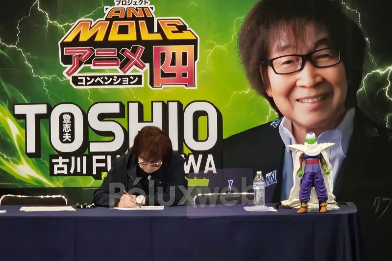 Toshio Furukawa en AniMole 4 El actor de voz japonés Toshio Furukawa sentado en su mesa de autógrafos firmando documentos, frente a un banner con su rostro y con una figura de Piccolo colocada sobre la mesa.