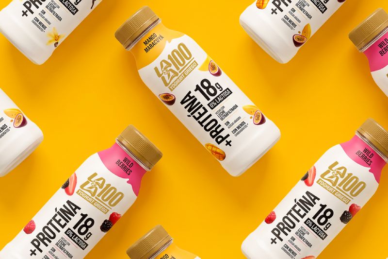 Nuevas botellas de Yogurt Lala 100 Griego con 18g de proteína Patrón diagonal de botellas de Yogurt Lala 100 Griego en fondo amarillo, mostrando variedades de sabores como Vainilla, Mango Maracuyá y Wild Berries con la etiqueta que destaca 18g de proteína y 0% lactosa.
