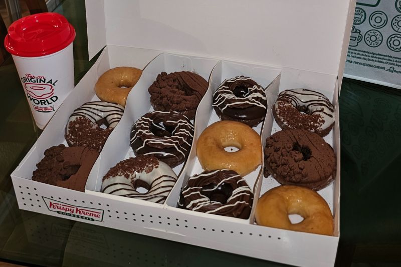 Caja abierta con una docena de donas surtidas colocada junto a un vaso de la marca sobre una superficie de vidrio.