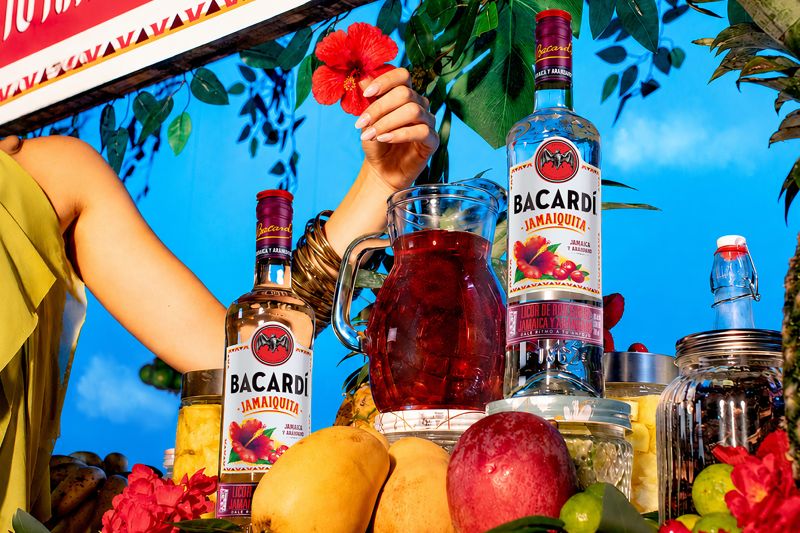 Botellas de Bacardí Jamaiquita y jarra preparada con hielos Botellas de cristal del ron saborizado Bacardí Jamaiquita expuestas en un puesto de frutas con flores rojas de jamaica, mangos y piñas. Al centro destaca una jarra de cristal con la bebida preparada de color rojo oscuro sobre un fondo de vegetación y cielo azul.