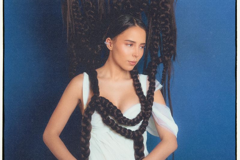 Retrato artístico de una mujer con un vestido blanco y una elaborada estructura de trenzas que rodean su cuerpo, sobre un fondo azul.