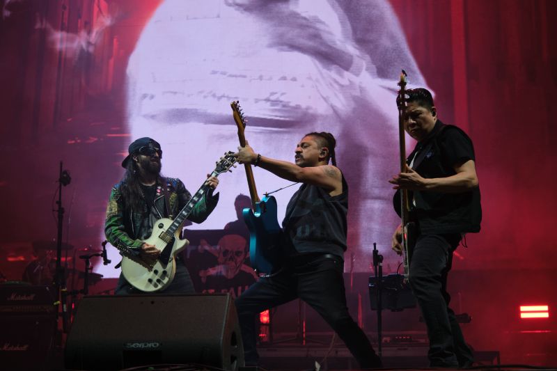 Tres músicos de Panteón Rococó tocan en un escenario iluminado con luces rojas y una gran pantalla al fondo, en la que se proyecta una imagen en blanco y negro, capturando la intensidad de un show en vivo.