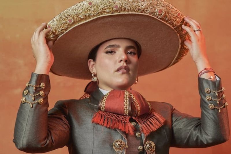 Fotografía de estudio de la cantante Camila Fernández posando con un elegante traje de chargris o gris oscuro con detalles y botonadura dorada de estilo tradicional. La artista porta un sombrero de charro de ala ancha en tono beige con bordados dorados, el cual sostiene con ambas manos a la altura de la cabeza, mientras luce una corbata de moño en tonos rojos y una expresión segura mirando directamente a la cámara sobre un fondo cálido difuminado.