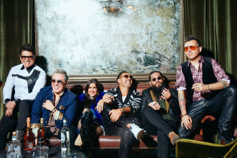 Integrantes de la banda Matute Integrantes de la banda Matute posando juntos en un sillón de piel dentro de un salón elegante, luciendo vestimenta de estilo moderno y gafas oscuras frente a una mesa con botellas.