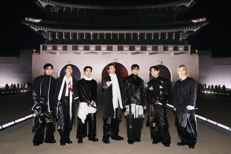 BTS en Gwanghwamun durante su regreso en vivo Siete integrantes posan frente a la puerta de Gwanghwamun iluminada de noche, vestidos con atuendos en blanco y negro y sosteniendo micrófonos.