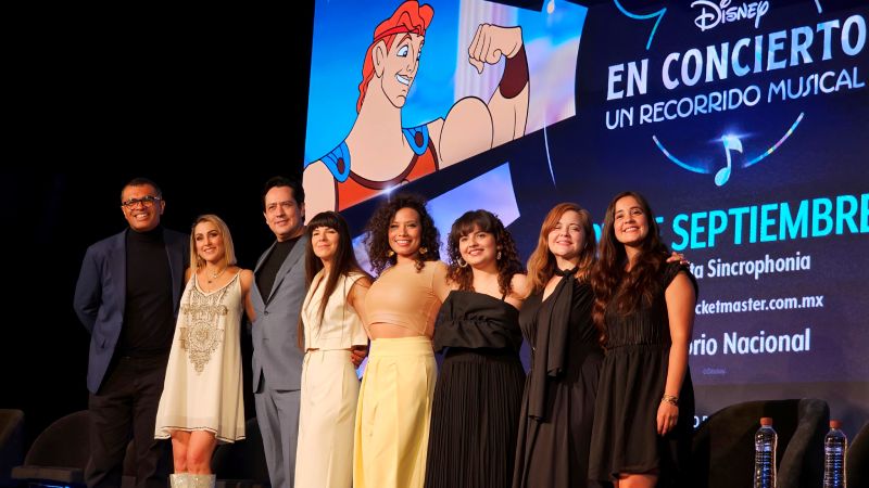 Presentación oficial — Disney en Concierto Siete integrantes del elenco y producción posan frente a una pantalla con arte promocional del concierto.