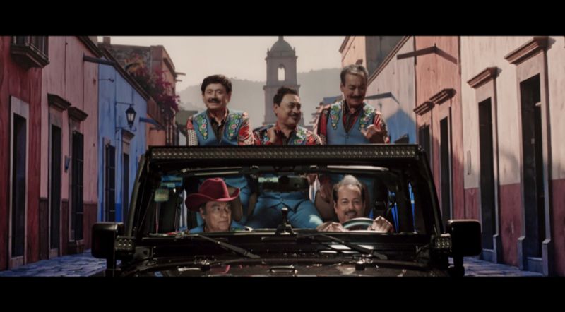Los Tigres del Norte presentan el video oficial de Mexico Sigue De Pie Los integrantes de Los Tigres del Norte viajan sonrientes a bordo de un vehículo todoterreno negro a través de una calle empedrada flanqueada por fachadas de colores y una torre de iglesia al fondo.