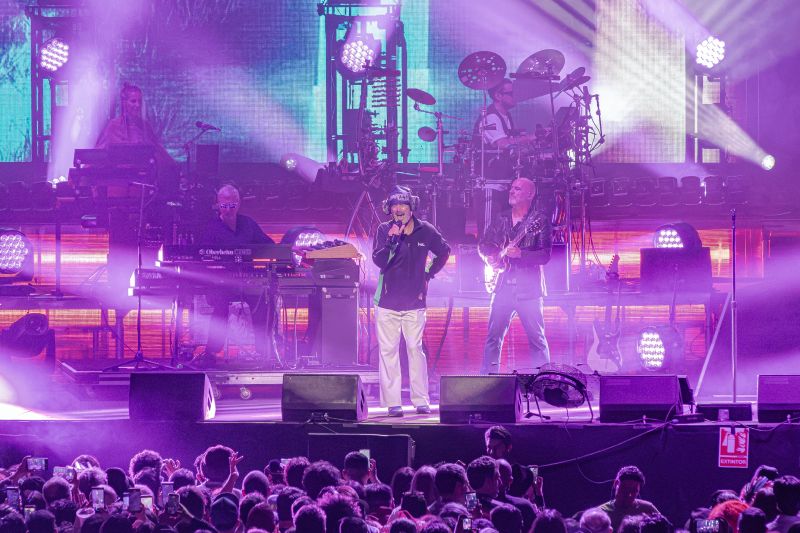 Jay Kay interactuando con la banda en vivo Jay Kay cantando al centro del escenario vistiendo sudadera y audífonos iluminados junto a los músicos de la banda bajo luces moradas.